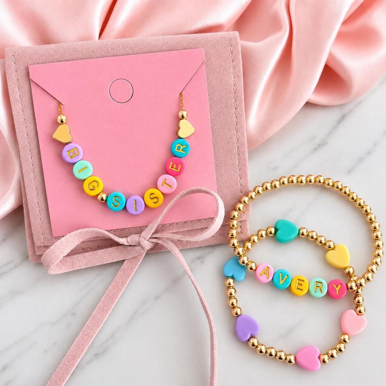 May include: A necklace and two bracelets featuring colourful round letter beads and heart-shaped beads on gold-tone chains. The necklace spells BIG SISTER with multi-coloured beads, while one bracelet spells AVERY. The jewellery is displayed on a pink card.