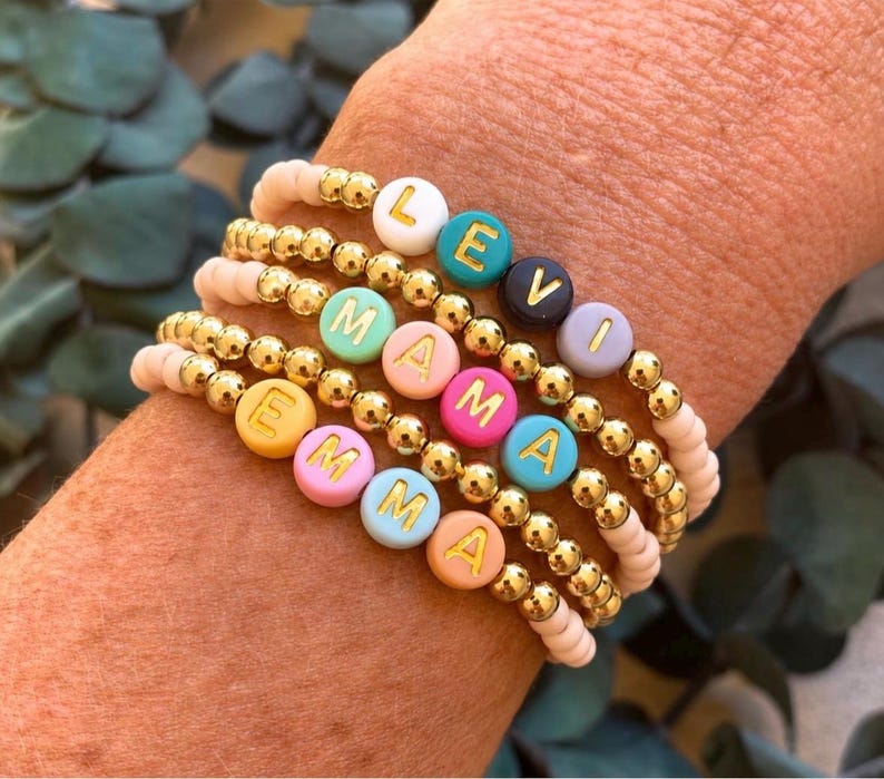 Pu&ograve; includere: Una pila di cinque braccialetti di perline con perline dorate metallizzate e perline opache color pastello. I braccialetti compongono i nomi LEVI, MAMA ed EMMA utilizzando perline rotonde con lettere bianche, verde acqua, blu scuro, lavanda, menta, rosa, giallo e pesca.