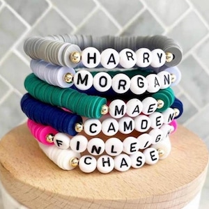 May include: Stack of colorful silicone bracelets with white beads spelling out the names "HARRY", "MORGAN", "MAE", "CAMDEN", "FINLEY", and "CHASE".