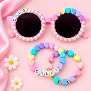May include: Pink sunglasses decorated with white faux pearls and the name CHLOE in white letter beads on the left lens, plus two elastic bracelets featuring colorful heart-shaped beads, gold-tone spacers, and the name CHLOE in white letter beads.