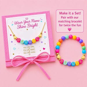 May include: A gold-tone chain necklace with colorful letter beads spelling BROOKE and gold heart charms on a pink display card with the text Wear Your Name Shine Bright, alongside a matching colorful beaded bracelet with the name BROOKE.