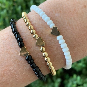 Può includere: Tre braccialetti con ciondoli a forma di cuore. I braccialetti sono neri, dorati e bianchi.
