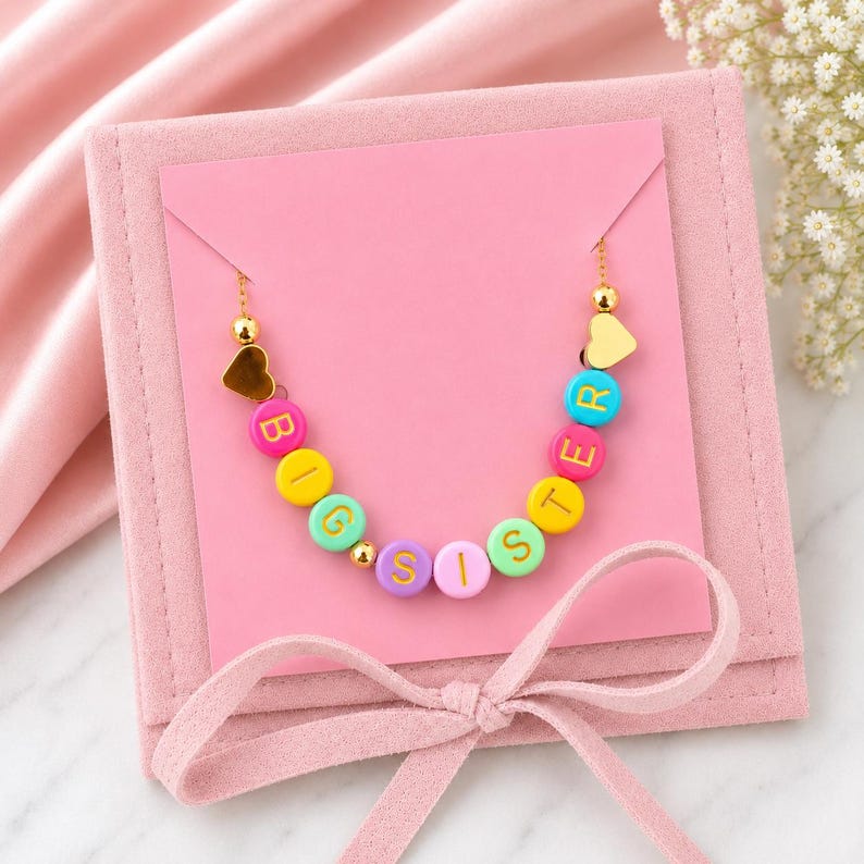 May include: A gold-toned chain necklace featuring colourful round beads that spell out BIG SISTER in yellow letters. The necklace includes small gold-toned heart charms at each end and is displayed on a square pink card with a ribbon tie.