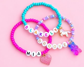 May include: Three beaded bracelets featuring white letter beads spelling the names BLAKELY, BROOKE, and MIA. The bracelets include colorful round beads, a gold-tone unicorn charm, a blue and purple gummy bear charm, and a pink glitter heart charm.
