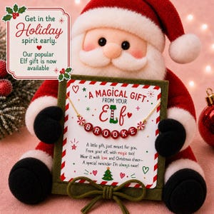 May include: A plush Santa Claus holding a square green gift box with a gold chain necklace featuring red and white beads spelling BROOKE. The card reads A MAGICAL GIFT FROM YOUR ELF and includes a poem about Christmas cheer and magic.