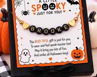 May include: A gold-toned chain bracelet with black letter beads spelling BROOKE, flanked by two gold heart charms. The bracelet is displayed on a white card with orange borders, featuring Halloween graphics and the text A LITTLE SPOOKY JUST FOR YOU.