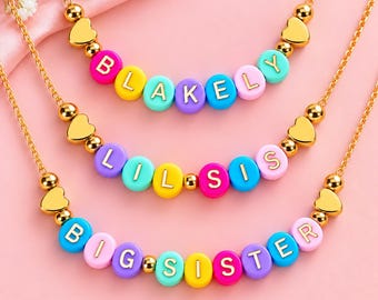 Puede incluir: Tres collares de cadena dorada con cuentas redondas de letras de colores y cuentas doradas en forma de corazón. Los collares muestran las palabras BLAKELY, LIL SIS y BIG SISTER en letras doradas sobre cuentas rosas, amarillas, azules y moradas.