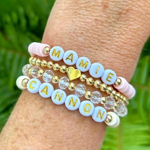 Può includere: Un set di braccialetti con i nomi "MAMIE" e "CANNON" scritti in lettere dorate su perline bianche. I braccialetti sono sovrapposti al polso di una persona e includono perline dorate e perline di cristallo trasparente.