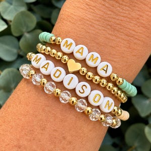 Puede incluir: Un conjunto de tres pulseras de cuentas. Una tiene cuentas verde menta y cuentas de letras blancas que forman la palabra MAMA. Otra tiene cuentas metálicas doradas y un dije de corazón dorado. La tercera consiste en cuentas de cristal transparente facetado y cuentas de letras blancas que forman MADISON.
