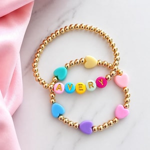 May include: Two stretch bracelets made of small gold-toned metallic beads. One bracelet features a series of pastel-coloured heart-shaped beads. The other bracelet features round beads spelling the name AVERY in gold letters, interspersed with heart beads.