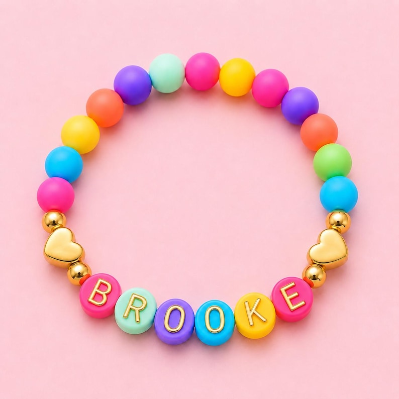 May include: A circular beaded bracelet featuring a vibrant array of multicolored matte spheres in shades of pink, purple, blue, yellow, orange, and green. The bracelet includes gold-toned heart charms and letter beads spelling BROOKE.