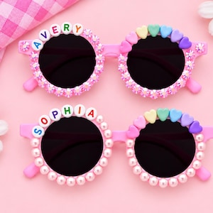 May include: Two pairs of pink sunglasses with round black lenses. The top pair features the name AVERY in colorful letter beads and pink crystals. The bottom pair features the name SOPHIA in colorful letter beads and white pearl-like beads.