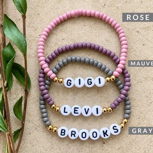 Pu&ograve; includere: Tre braccialetti di perline con perline rotonde nei colori rosa, malva e grigio. Ogni braccialetto include perline distanziatrici dorate e perline circolari bianche con lettere nere che compongono i nomi GIGI, LEVI e BROOKS.