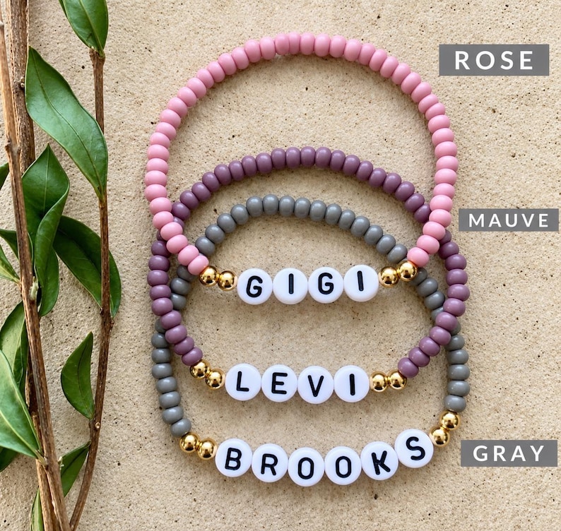 Pu&ograve; includere: Tre braccialetti di perline con perline rotonde nei colori rosa, malva e grigio. Ogni braccialetto include perline distanziatrici dorate e perline circolari bianche con lettere nere che compongono i nomi GIGI, LEVI e BROOKS.