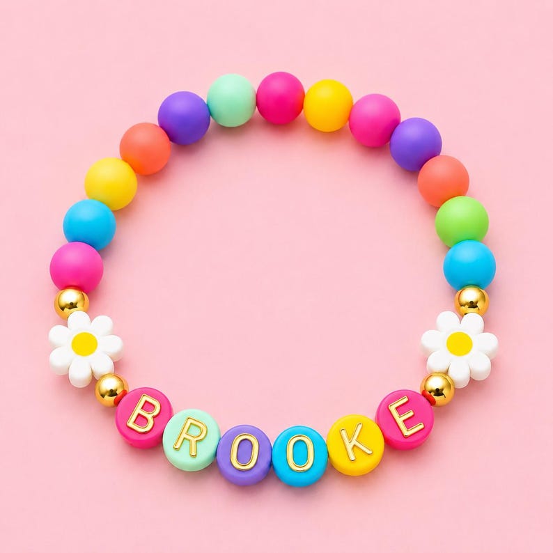May include: A circular beaded bracelet featuring a variety of matte-finish, multi-colored spherical beads in shades of pink, yellow, blue, orange, purple, and green. Two white daisy-shaped beads with yellow centers are included, along with gold-toned spacer beads and letter beads spelling out the name BROOKE in gold lettering.