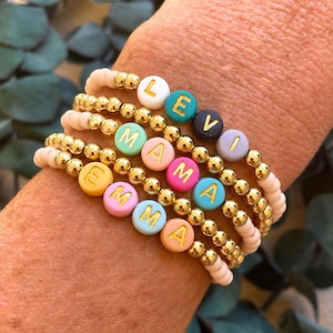 May include: A stack of five beaded bracelets featuring metallic gold beads and matte pastel-coloured letter beads. The bracelets spell out the names LEVI, MAMA, and EMMA using gold-coloured letters on round beads in shades of white, teal, navy, lavender, mint, pink, yellow, blue, and peach.