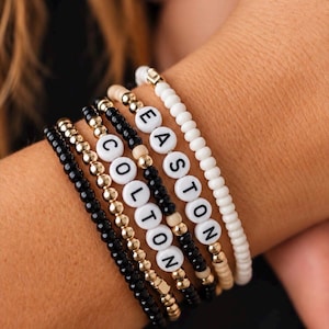 Pu&ograve; includere: Una pila di cinque braccialetti di perline con piccole perline nere, dorate e color crema. Due braccialetti includono perline circolari bianche con lettere nere che compongono i nomi EASTON e COLTON in una disposizione verticale.