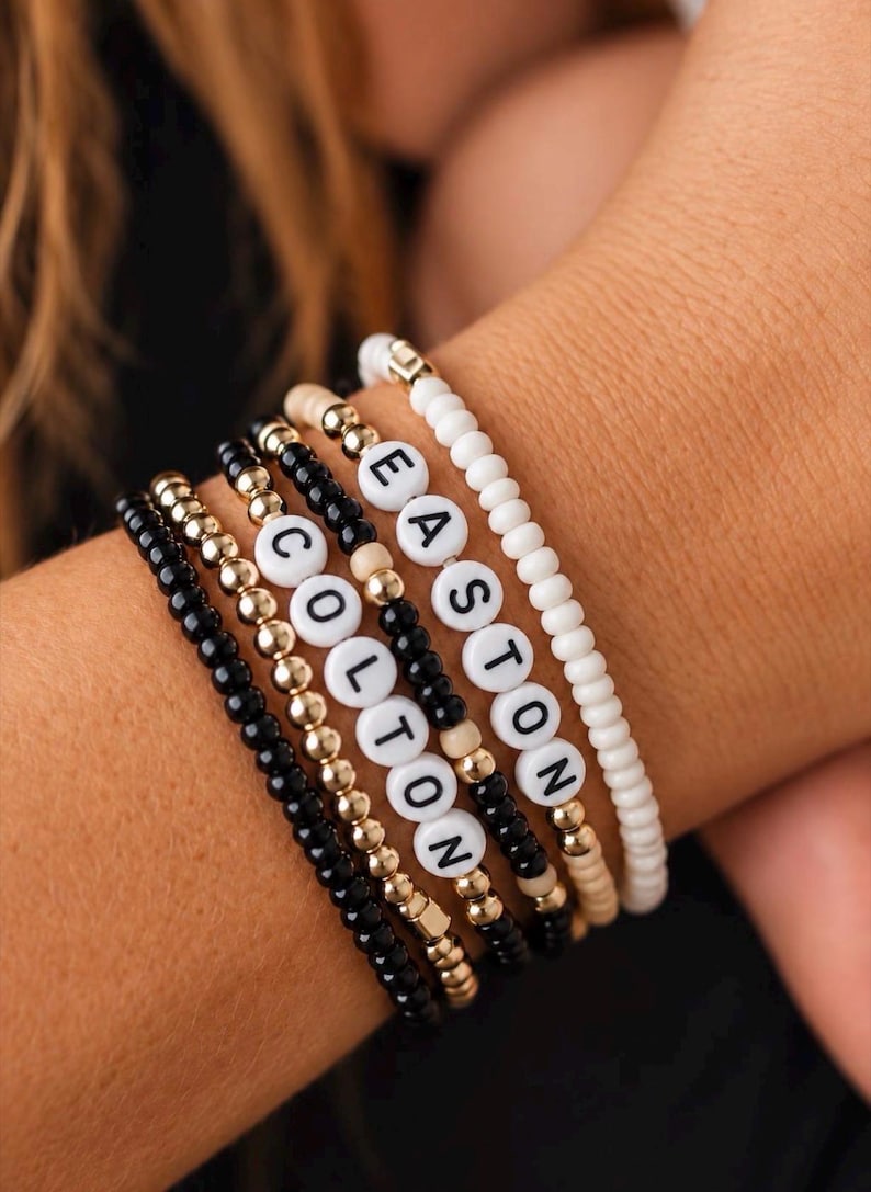 Pu&ograve; includere: Una pila di cinque braccialetti di perline con piccole perline nere, dorate e color crema. Due braccialetti includono perline circolari bianche con lettere nere che compongono i nomi EASTON e COLTON in una disposizione verticale.