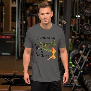 May include: A dark gray t-shirt with a graphic of a green and brown Grumman F6F Hellcat airplane with a pin-up girl in a yellow dress. The text "Grumman F6F Hellcat" is printed on the shirt.