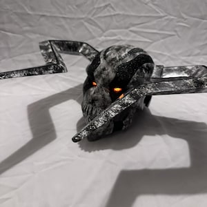 May include: A skull-shaped helmet featuring a marbled black and white finish with large, angular, lightning-bolt-shaped horns extending from the sides. The eye sockets are illuminated with glowing orange light, and the surface has a textured appearance.