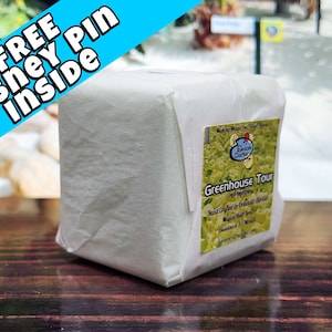 May include: A white cube-shaped package wrapped in white paper with a label that reads "Greenhouse Tour" and "Hand Crafted in Orlando, Florida". The package is sitting on a wooden surface.
