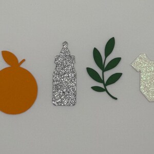 May include: A collection of paper cutouts including a quarter, an orange, a glittery bottle, a green leaf sprig, a glittery baby onesie, and a slice of orange. The objects are arranged in a row on a white surface.