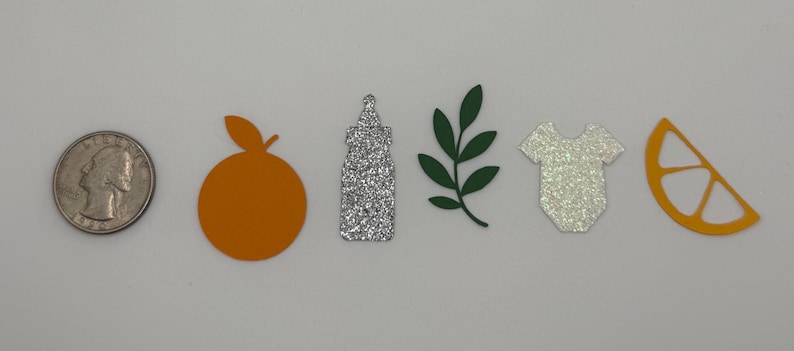 May include: A collection of paper cutouts including a quarter, an orange, a glittery bottle, a green leaf sprig, a glittery baby onesie, and a slice of orange. The objects are arranged in a row on a white surface.