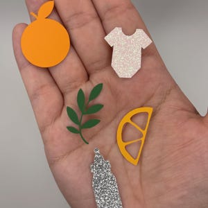 May include: A collection of colorful paper cutouts, including an orange, a baby onesie, a lemon slice, a green leaf sprig, and a glittery bottle. The orange is bright orange, the onesie is white with glitter, and the lemon slice is yellow.