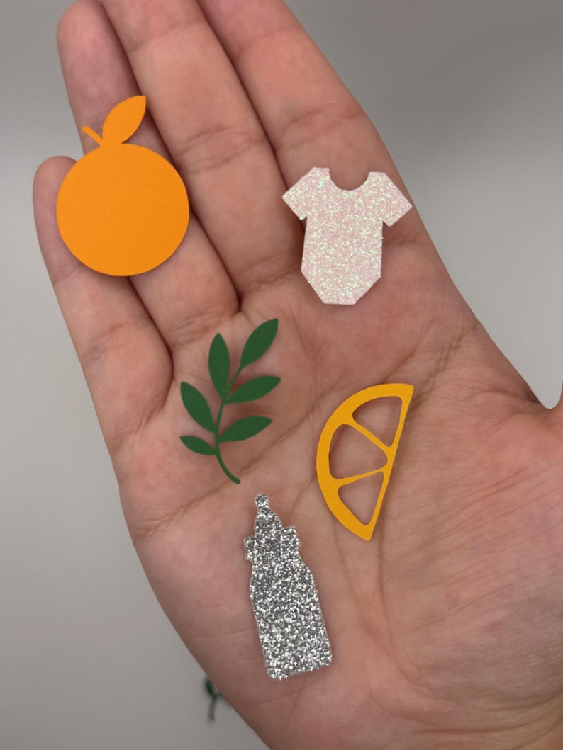 May include: A collection of colorful paper cutouts, including an orange, a baby onesie, a lemon slice, a green leaf sprig, and a glittery bottle. The orange is bright orange, the onesie is white with glitter, and the lemon slice is yellow.