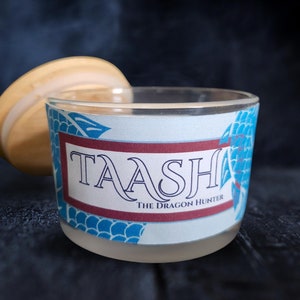 May include: A glass candle jar with a blue and white label that says "TAASH THE DRAGON HUNTER". The jar has a wooden lid.