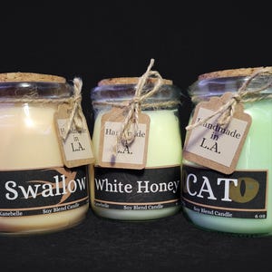 Może przedstawiać: Trzy szklane słoiki z korkowymi pokrywkami i sznurkowymi kokardkami. Słoiki zawierają świece sojowe w zapachach Swallow, White Honey i CAT. Każdy słoik ma brązową papierową etykietę z napisem "Handmade in L.A."