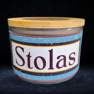 May include: A glass candle jar with a bamboo lid. The jar has a blue and gold label with the word "Stolas" printed in red.