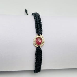 May include: A black woven cord bracelet featuring a central gold-toned circular charm with a textured, hammered finish. The charm holds a single, small, oval-shaped red gemstone in the center, secured by a metal setting.