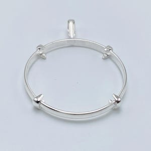 May include: A circular silver pendant setting featuring a smooth, thin metal band with four evenly spaced prongs extending inward to secure a stone. The pendant includes a small, vertical bail at the top for attachment to a chain or necklace.