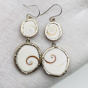 May include: Dangle earrings with white spiral shell designs set in silver-coloured metal. The earrings have a two-tiered design with a small, round shell at the top and a larger, round shell below, connected by a small metal loop.