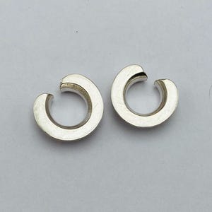 Może przedstawiać: Dwie srebrne nausznice. Każda nausznica ma kształt litery C z małym otworem. Kolczyki są gładkie i błyszczące, odbijają światło. Tło jest jasnoszare.