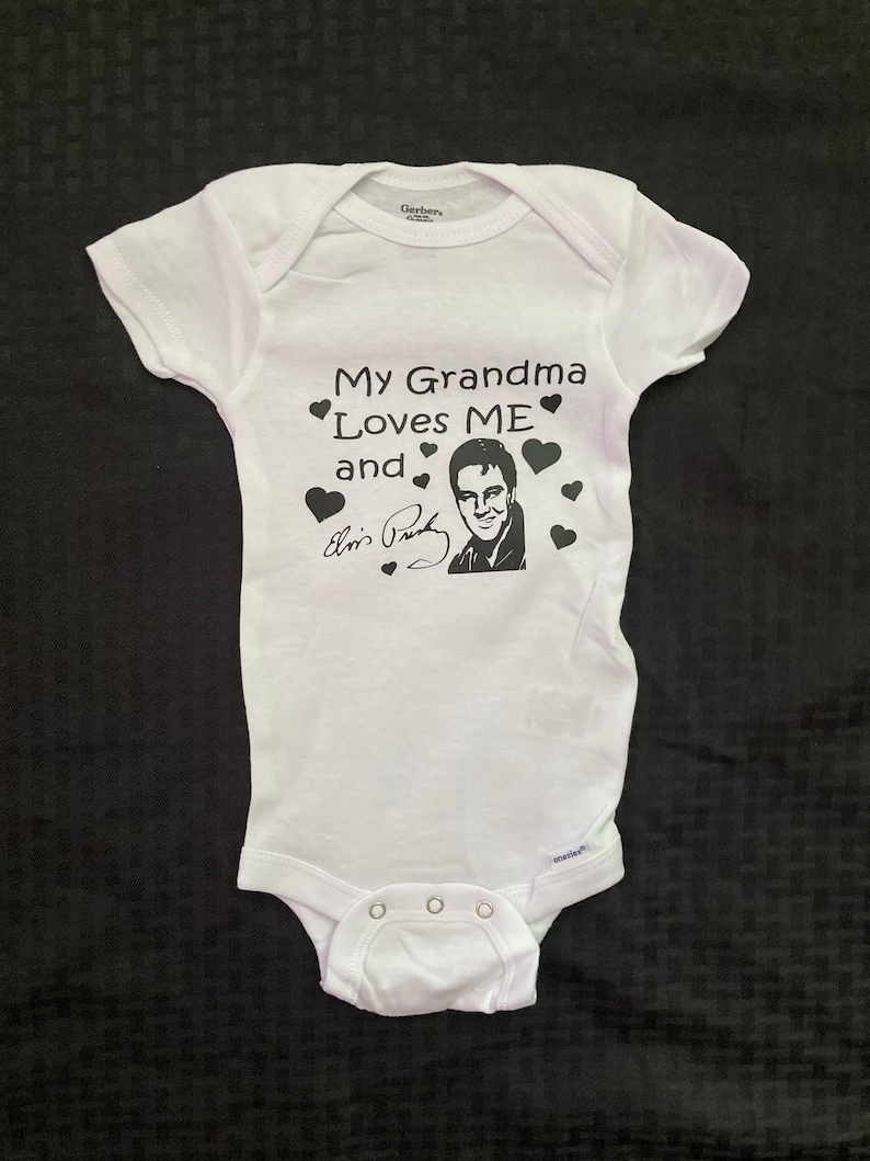 May include: White baby onesie with black text that reads "My Grandma Loves ME and" with a black and white image of Elvis Presley and his signature.