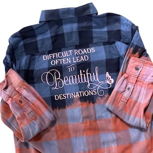 Może przedstawiać: Koszula flanelowa w kratę niebiesko-pomarańczową z napisem "Difficult roads often lead to beautiful destinations" w różowych literach na plecach.