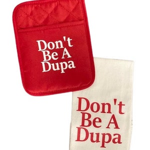 May include: A red quilted potholder with a hanging loop and a white kitchen towel. Both items feature the text Don't Be A Dupa printed in a bold font. The potholder has white text on a red background, while the towel has red text on a white background.