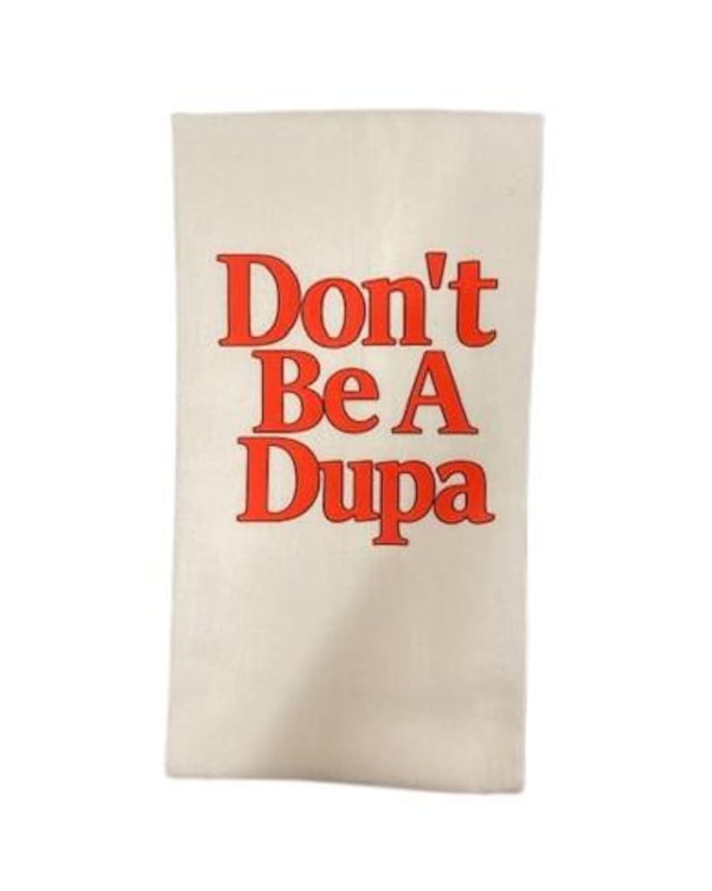 May include: A rectangular off-white kitchen towel featuring the text Don't Be A Dupa printed in a bold, bright orange-red serif font. The fabric has a visible woven texture, and the lettering is centered vertically and horizontally on the towel.