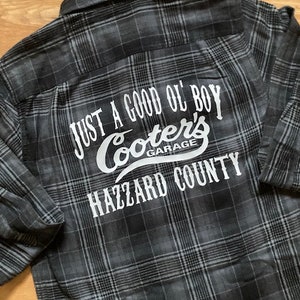 May include: A black and gray plaid flannel shirt with the text "Just a good ol' boy Cooter's Garage Hazzard County" printed on the back.