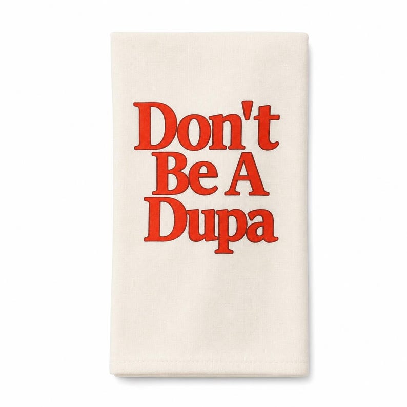May include: A rectangular off-white kitchen towel featuring the text Don't Be A Dupa printed in a bold, bright red serif font. The fabric has a smooth, woven texture and the lettering is centered on the front of the folded textile item.