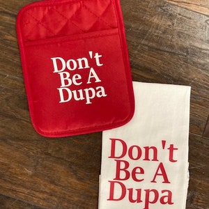 May include: A red quilted oven mitt with a hanging loop and a white kitchen towel. Both items feature the white and red text Don't Be A Dupa printed in a bold serif font. The oven mitt has a rectangular shape with rounded corners and a pocket.
