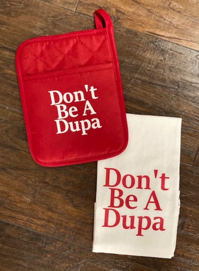 May include: A red quilted oven mitt with a hanging loop and a white kitchen towel. Both items feature the white and red text Don't Be A Dupa printed in a bold serif font. The oven mitt has a rectangular shape with rounded corners and a pocket.