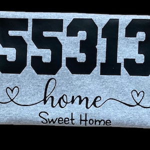 May include: A gray t-shirt with black text that reads "55313 home Sweet Home" with two hearts on either side of the word "home".