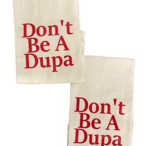 May include: Two rectangular off-white kitchen towels featuring the red text Don't Be A Dupa printed in a bold serif font. The towels are positioned slightly offset from each other against a plain white background, displaying the same text.