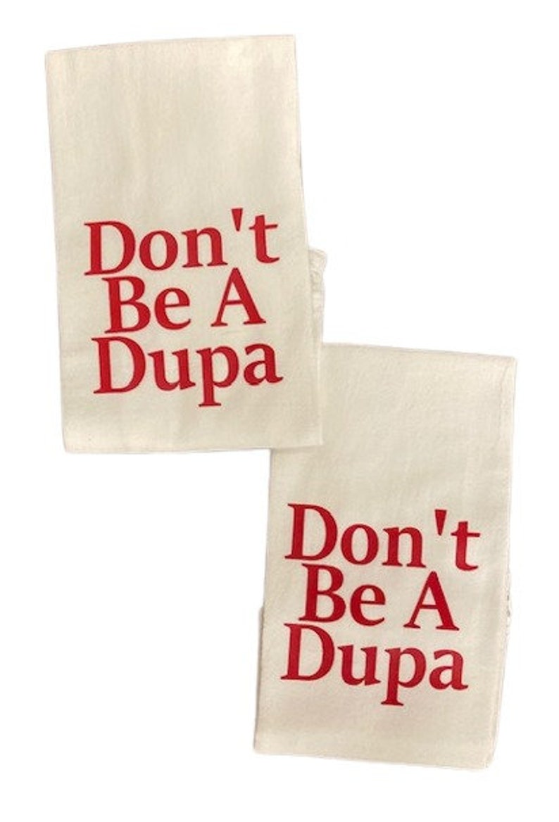 May include: Two rectangular off-white kitchen towels featuring the red text Don't Be A Dupa printed in a bold serif font. The towels are positioned slightly offset from each other against a plain white background, displaying the same text.