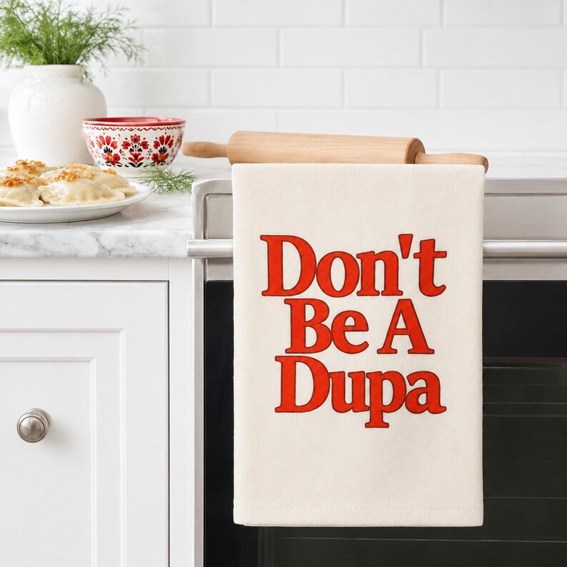May include: A cream-colored kitchen towel hanging over a wooden rolling pin, featuring the text Don't Be A Dupa printed in bold, red, sans-serif capital letters. The towel is made of fabric and displays a clean, rectangular design.