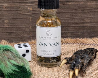 May include: A clear glass bottle with a black cap containing liquid and herbs, labeled Conjured Moon Van Van Conjure Oil 0.5 fl oz / 15 ml. Beside it sits a white six-sided die, a tuft of green fur, and a dark, textured animal claw with sharp nails.