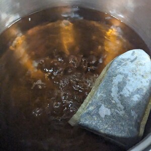 May include: A stainless steel pot filled with boiling, dark brown liquid. A tea bag is partially submerged in the water. Bubbles rise to the surface, indicating the water is hot. The pot sits on a red stovetop.